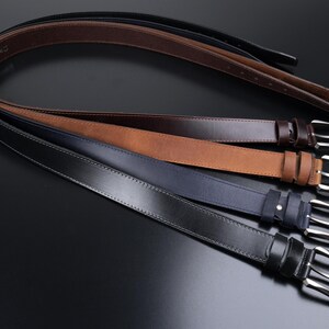 May include: A collection of four leather belts arranged in a stack, featuring a black, a dark blue, a tan, and a dark brown belt. Each belt is equipped with a silver-toned metal pin buckle and a matching leather keeper loop for secure fastening.