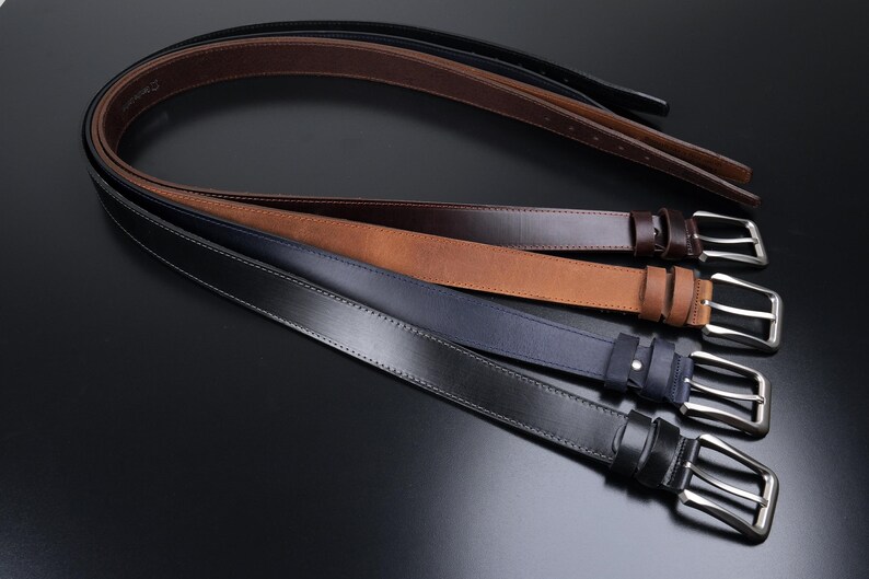May include: A collection of four leather belts arranged in a stack, featuring a black, a dark blue, a tan, and a dark brown belt. Each belt is equipped with a silver-toned metal pin buckle and a matching leather keeper loop for secure fastening.