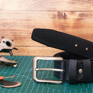May include: A dark navy blue leather belt featuring a silver-toned metal buckle and matching belt loops. The belt is coiled and displays visible stitching along the edges. A metal hole punch tool and a small seam ripper are positioned nearby.