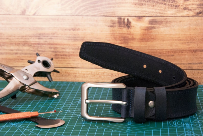 May include: A dark navy blue leather belt featuring a silver-toned metal buckle and matching belt loops. The belt is coiled and displays visible stitching along the edges. A metal hole punch tool and a small seam ripper are positioned nearby.