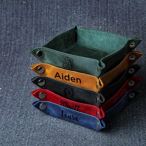 May include: A stack of five leather valet trays in different colors, including green, yellow, black, red, and blue. Each tray has a personalized name, including Aiden, Wyatt, and Laura.