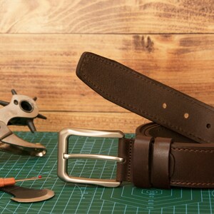 May include: A dark brown leather belt featuring a rectangular silver-toned metal buckle and matching leather keepers. The belt has visible stitching along the edges and several punched holes for adjustment, presented alongside a metal hole punch tool.