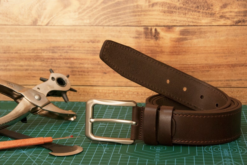 May include: A dark brown leather belt featuring a rectangular silver-toned metal buckle and matching leather keepers. The belt has visible stitching along the edges and several punched holes for adjustment, presented alongside a metal hole punch tool.