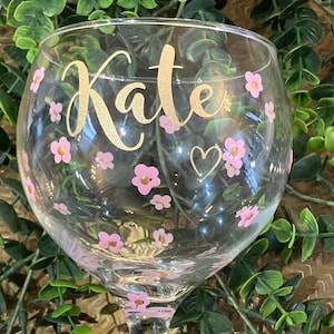 May include: A clear glass balloon gin glass featuring the name Kate in gold cursive lettering. The glass is decorated with small pink flowers with gold centres and a small gold heart outline scattered across the surface of the bowl.