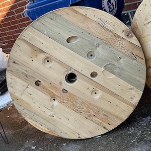 May include: A large wooden spool with a center hole and multiple holes around the edge. The spool is made of different types of wood, including light brown, dark brown, and gray. The spool has a weathered look and is likely used for storing cable or wire.
