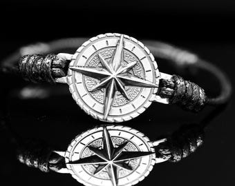 May include: A silver compass rose bracelet with a black rope band. The compass rose features a detailed star design. The bracelet is set against a black background, creating a striking contrast.