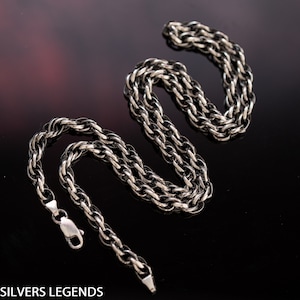 May include: A silver chain necklace with a twisted rope design. The chain is laid out in a figure eight shape on a black surface.