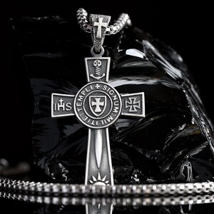 May include: A silver cross pendant with a sword design and the words "IHS TEMPLI MILITIE SIGNUM" around the center. The cross is on a black chain.