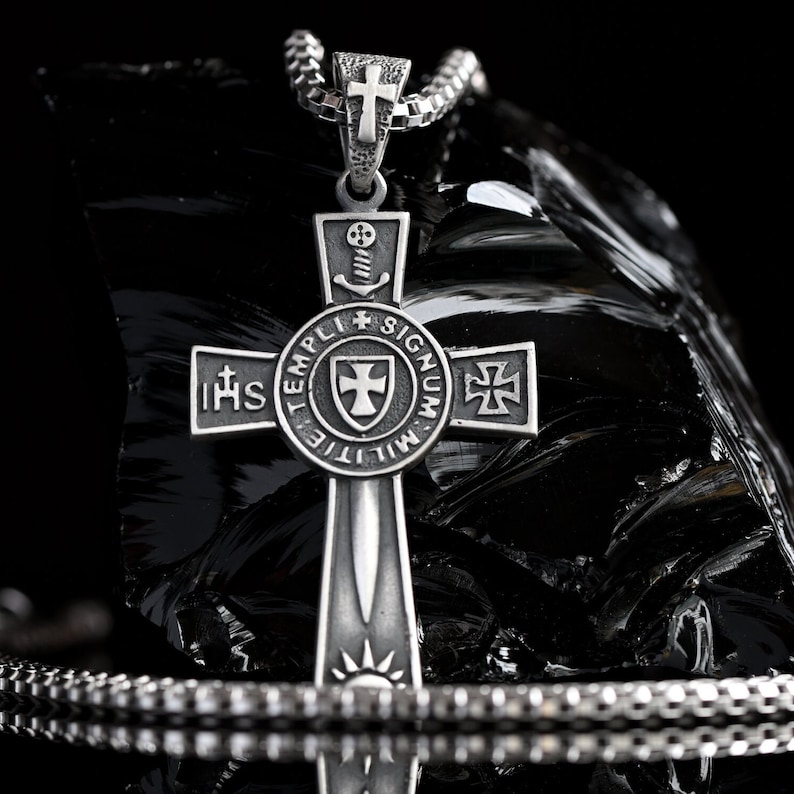 May include: A silver cross pendant with a sword design and the words "IHS TEMPLI MILITIE SIGNUM" around the center. The cross is on a black chain.