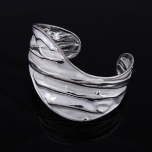 May include: A silver cuff bracelet with a wavy, textured design. The bracelet is open and has a wide, curved shape.