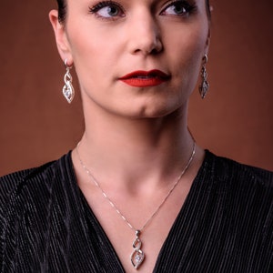 May include: A silver jewelry set featuring a necklace and matching earrings. The pendant and earrings have a swirling design with a central clear gemstone. The woman in the image is wearing a black top.