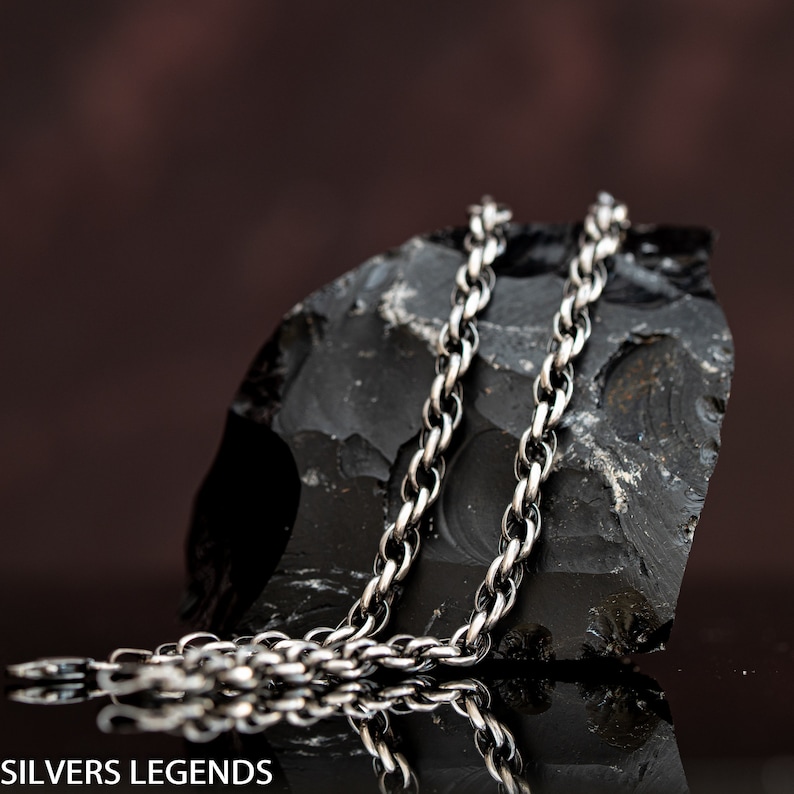 May include: A silver chain necklace with a rope chain design. The chain is lying on a black rock.
