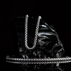 May include: A silver chain necklace with a box chain pattern, displayed on a black rock. The chain is laying on a black surface, reflecting the light.