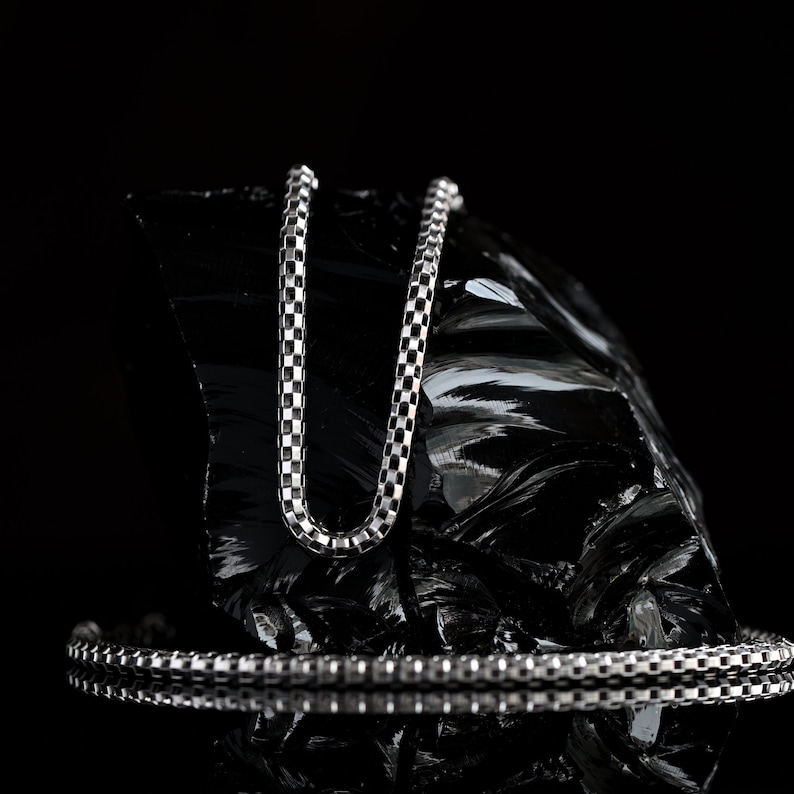 May include: A silver chain necklace with a box chain pattern, displayed on a black rock. The chain is laying on a black surface, reflecting the light.