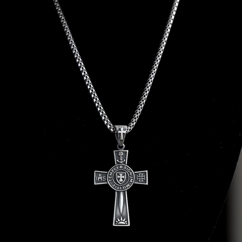May include: Silver chain necklace with a silver cross pendant. The cross features a circular design with the words "TEMPLI SIGNUM" and "IHHS" around the edge. The center of the cross has a shield with a sword and a cross.
