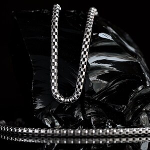 May include: A silver chain necklace with a box chain design. The chain is lying on a black, textured surface.
