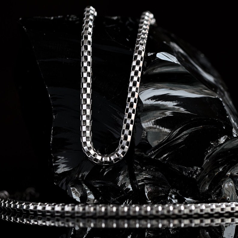 May include: A silver chain necklace with a box chain design. The chain is lying on a black, textured surface.