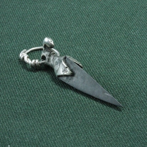 May include: A silver-toned pendant with a dagger-shaped blade and a detailed figure at the top. The pendant has a loop for a chain and is set against a dark green fabric background.