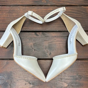 Puede incluir: Un par de tacones de novia de sat&eacute;n blanco con ribete de perlas y tac&oacute;n de bloque. Los tacones est&aacute;n dispuestos en forma de coraz&oacute;n sobre una superficie de madera.