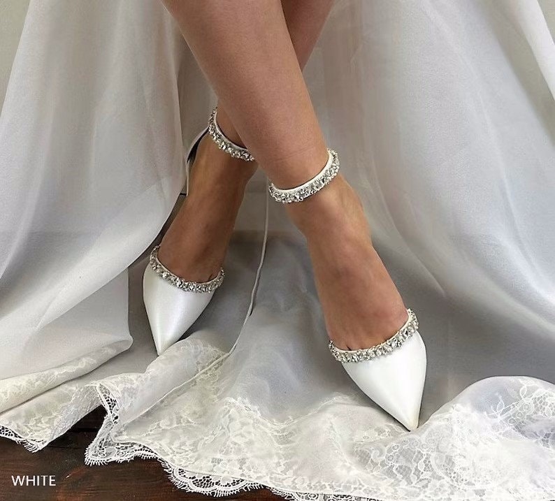 May include: White satin bridal shoes with a pointed toe and ankle strap adorned with sparkling rhinestones. The shoes are on a white lace background.