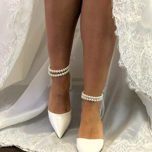 May include: A pair of white satin bridal shoes with pointed toes. The shoes have pearl ankle straps.