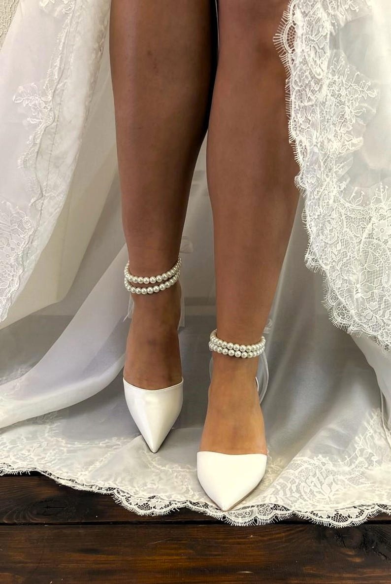 May include: A pair of white satin bridal shoes with pointed toes. The shoes have pearl ankle straps.