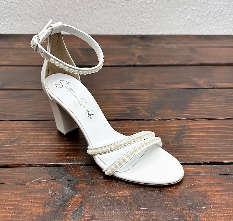 Puede incluir: Sandalias blancas de tac&oacute;n con adorno de perlas en las tiras y la tira del tobillo. Las sandalias tienen un tac&oacute;n de bloque y est&aacute;n hechas de cuero. El nombre de la marca "S-T-E-F-A-N-O-S-C-H-A-L-L" est&aacute; impreso en la plantilla.