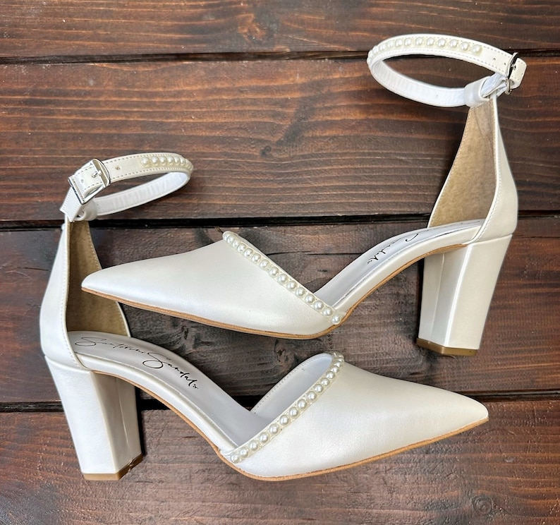 Puede incluir: Un par de zapatos de tac&oacute;n de sat&eacute;n blanco con adornos de perlas en las tiras y los tacones. Los zapatos tienen una punta puntiaguda y un tac&oacute;n de bloque.