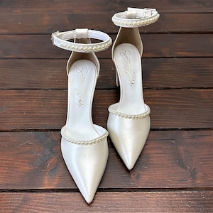 Puede incluir: Un par de tacones de novia de sat&eacute;n blanco con punta puntiaguda y adornos de perlas en la tira del tobillo y la punta. Los tacones est&aacute;n etiquetados como "Something Special".