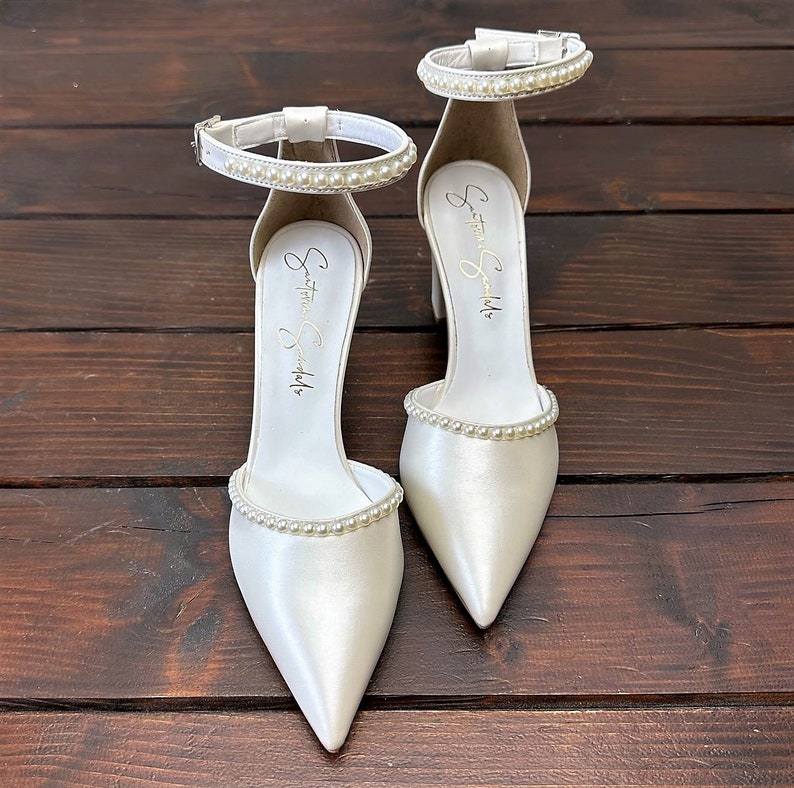 Puede incluir: Un par de tacones de novia de sat&eacute;n blanco con punta puntiaguda y adornos de perlas en la tira del tobillo y la punta. Los tacones est&aacute;n etiquetados como "Something Special".