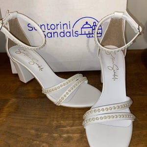 Puede incluir: Sandalias blancas de tac&oacute;n con adornos de perlas. Las sandalias tienen un tac&oacute;n grueso y una correa de tobillo. El nombre de la marca "Santorini Sandals" est&aacute; impreso en la caja.