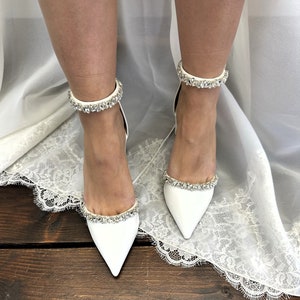 May include: White satin bridal shoes with a pointed toe and ankle strap adorned with crystal embellishments.