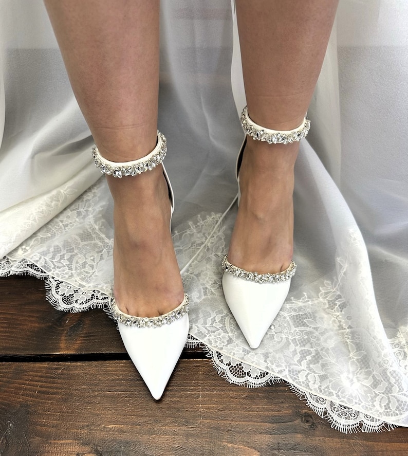 May include: White satin bridal shoes with a pointed toe and ankle strap adorned with crystal embellishments.