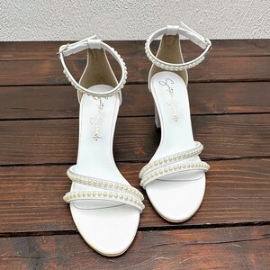 Puede incluir: Sandalias blancas de tac&oacute;n con adornos de perlas y tiras de tobillo. Las sandalias tienen el texto "Something Special" escrito en el interior de las tiras.