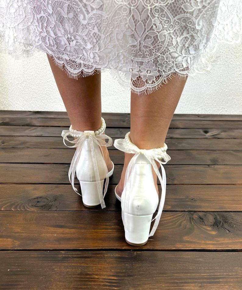 May include: White satin heeled sandals with a pearl ankle strap and a white satin bow. The sandals have a block heel.