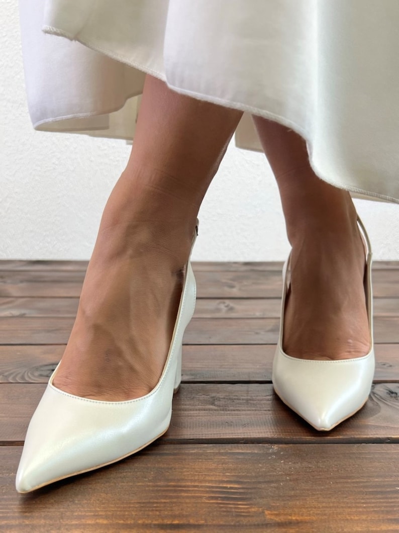 May include: A pair of white satin heeled shoes with a pointed toe and an ankle strap. The shoes are on a wooden floor.