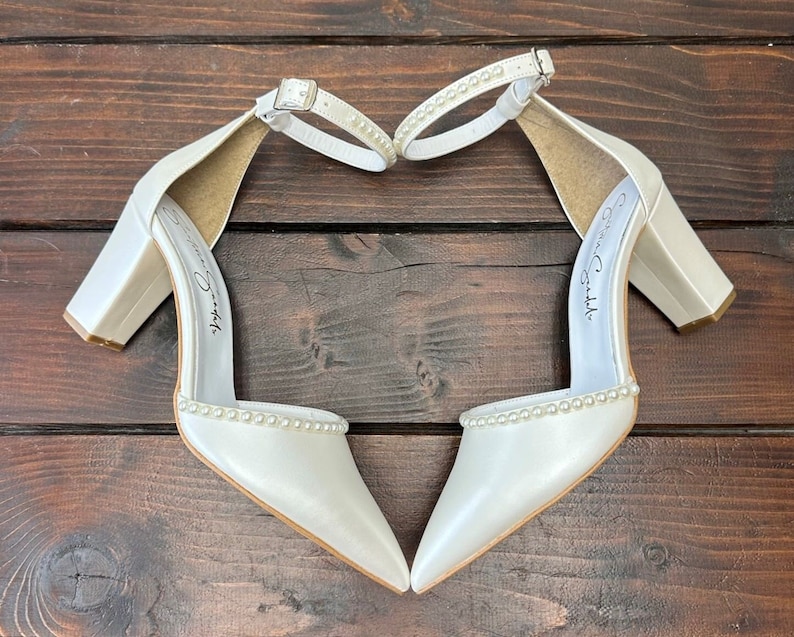 Puede incluir: Un par de tacones de novia de sat&eacute;n blanco con ribete de perlas y tac&oacute;n de bloque. Los tacones est&aacute;n dispuestos en forma de coraz&oacute;n sobre una superficie de madera.