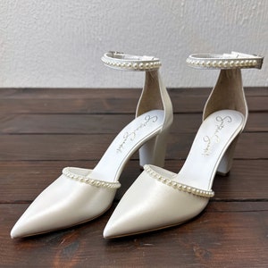 Puede incluir: Un par de zapatos de novia de sat&eacute;n blanco con adornos de perlas en las tiras y los dedos. Los zapatos tienen un tac&oacute;n de bloque y est&aacute;n firmados "Sophia Webster" en el interior.