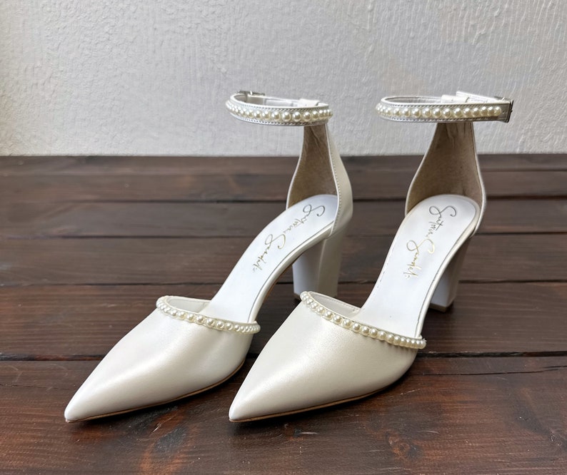 Puede incluir: Un par de zapatos de novia de sat&eacute;n blanco con adornos de perlas en las tiras y los dedos. Los zapatos tienen un tac&oacute;n de bloque y est&aacute;n firmados "Sophia Webster" en el interior.