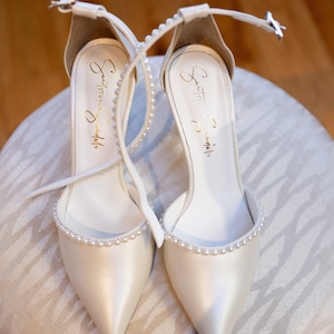 Puede incluir: Un par de zapatos de novia de sat&eacute;n blanco con adornos de perlas. Los zapatos tienen una punta puntiaguda y una delicada correa de tobillo. La inscripci&oacute;n "Something  Something" es visible en el interior de cada zapato.