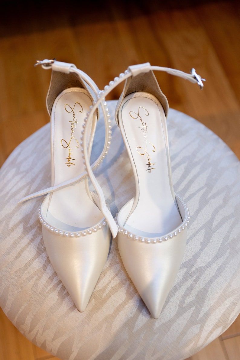 Puede incluir: Un par de zapatos de novia de sat&eacute;n blanco con adornos de perlas. Los zapatos tienen una punta puntiaguda y una delicada correa de tobillo. La inscripci&oacute;n "Something  Something" es visible en el interior de cada zapato.