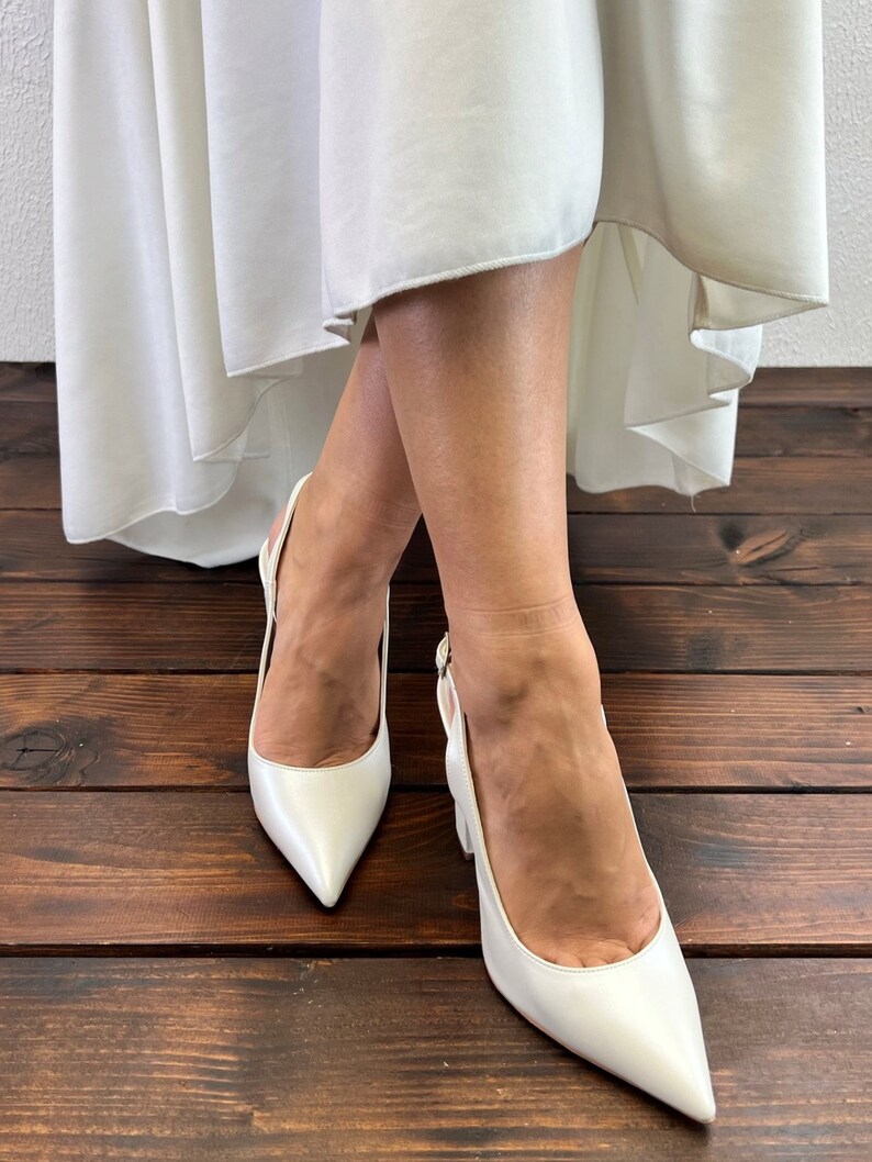 May include: A pair of white satin heeled shoes with a pointed toe and slingback strap. The shoes are worn by a person standing on a wooden floor.