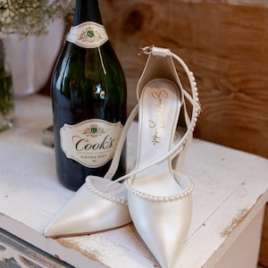 Puede incluir: Un par de zapatos de novia de sat&eacute;n blanco con adornos de perlas, una botella de champ&aacute;n Cook's Extra Dry de California y una mesita de noche de madera blanca.