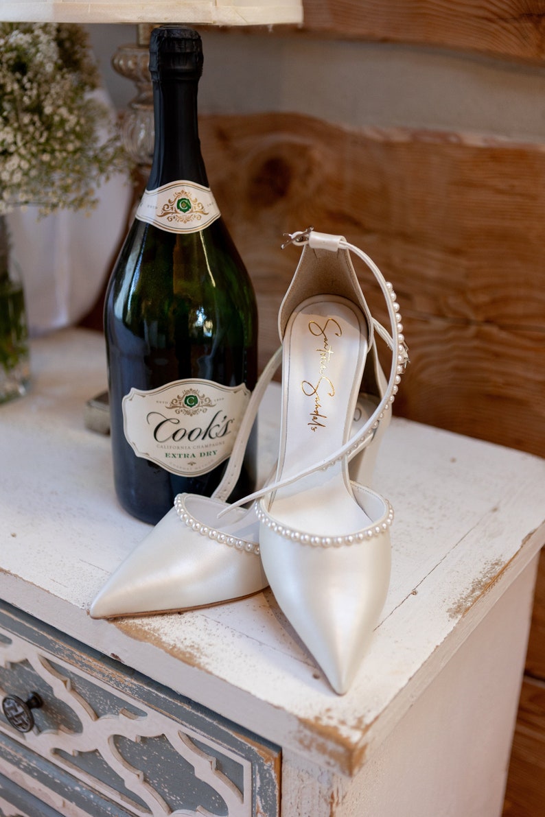 Puede incluir: Un par de zapatos de novia de sat&eacute;n blanco con adornos de perlas, una botella de champ&aacute;n Cook's Extra Dry de California y una mesita de noche de madera blanca.