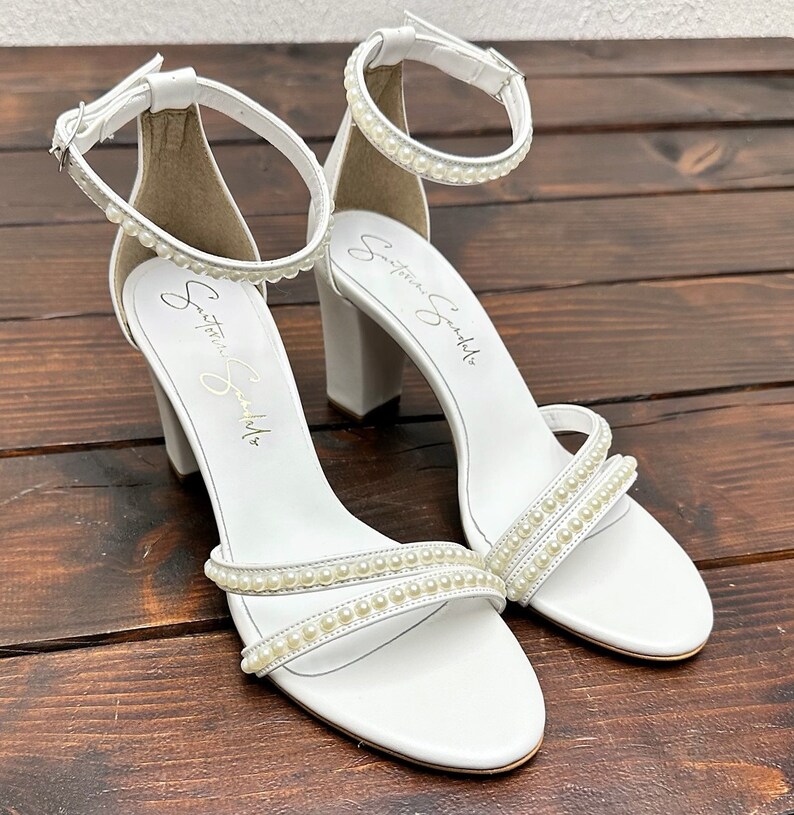 Puede incluir: Sandalias blancas de tac&oacute;n con adornos de perlas. Las sandalias tienen un tac&oacute;n grueso y una correa de tobillo. El nombre de la marca "Signature Sandals" est&aacute; escrito en la suela.
