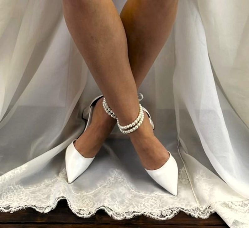 May include: White satin bridal shoes with a pointed toe and a pearl ankle bracelet. The shoes are worn by a person sitting on a white satin fabric.