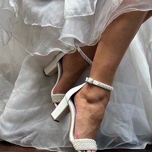 Puede incluir: Tacones de novia blancos con un adorno de perlas en la tira del tobillo y la tira del dedo del pie. Los tacones est&aacute;n sobre un suelo de madera.