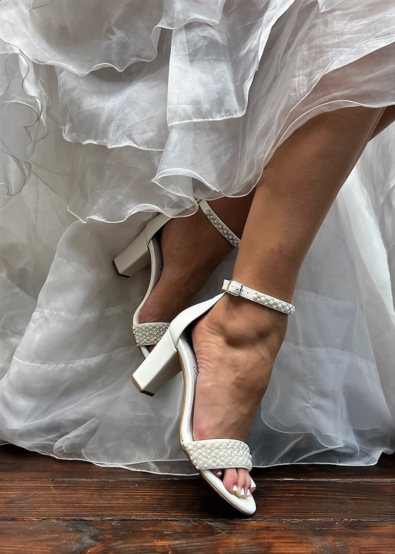 Puede incluir: Tacones de novia blancos con un adorno de perlas en la tira del tobillo y la tira del dedo del pie. Los tacones est&aacute;n sobre un suelo de madera.