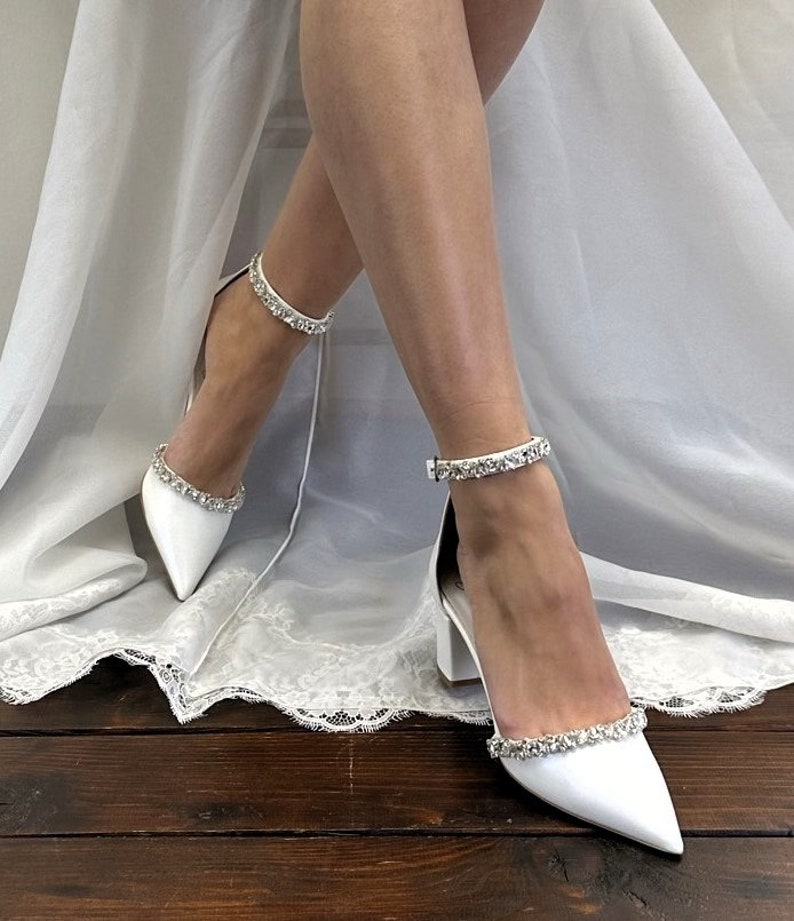 May include: White satin bridal shoes with a pointed toe and a block heel. The shoes have a crystal embellishment around the ankle strap and the toe.