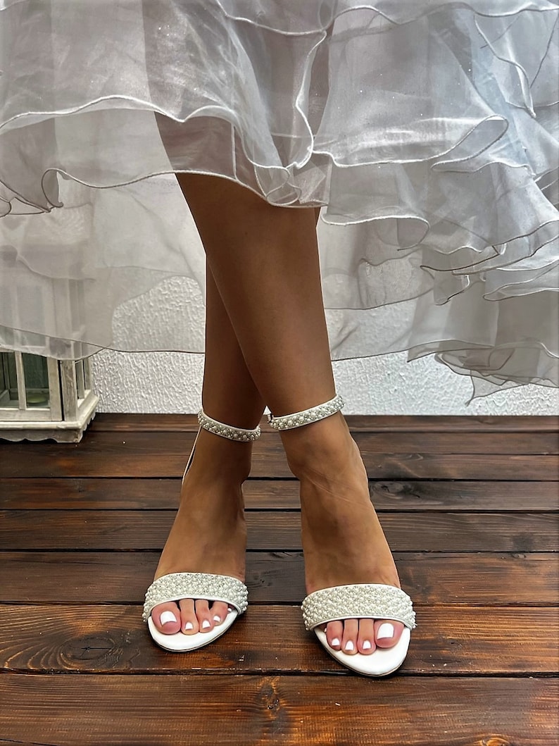 Puede incluir: Tacones de novia blancos con adornos de perlas en las tiras y la punta. Los tacones est&aacute;n sobre un suelo de madera.
